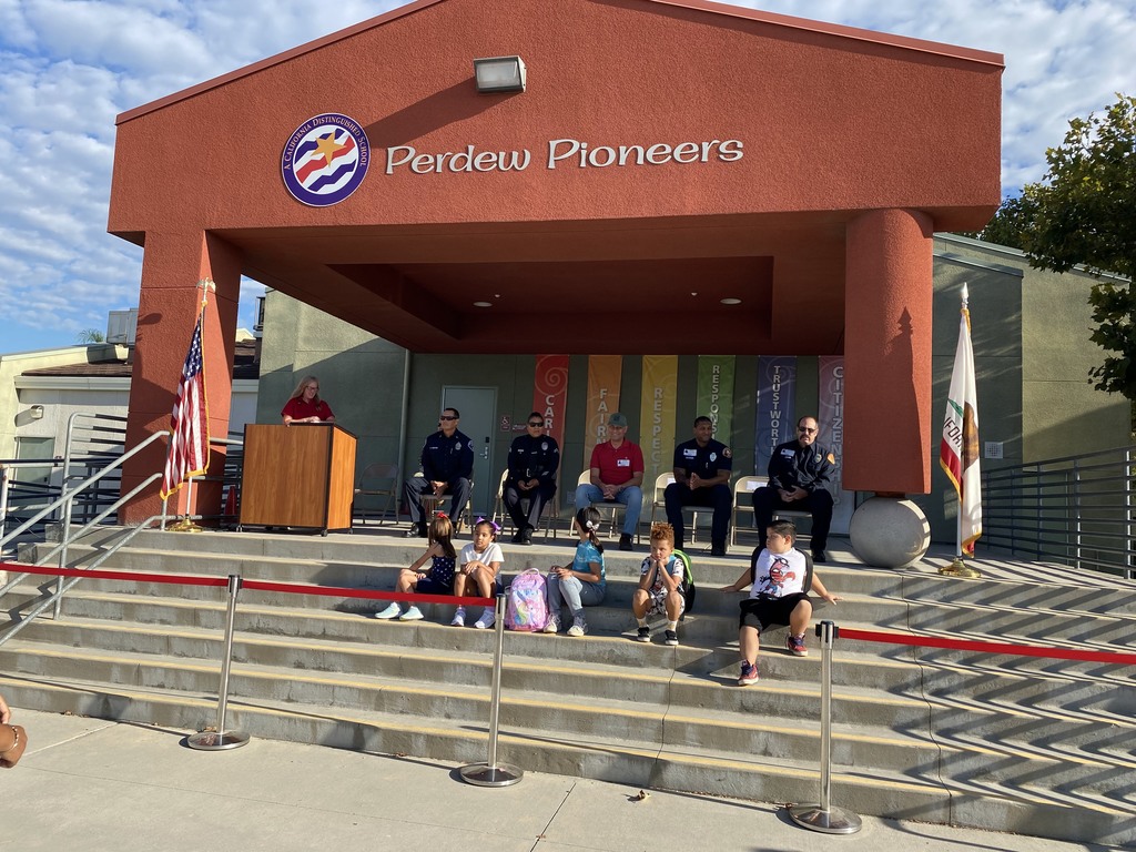 Firefighter, police officers, military personnel gathered on Perdew Elementary outdoor stage with students sitting on steps and principal at the podium